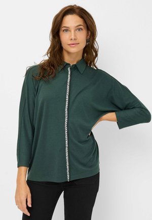 Chemise vert foncé en tissu doux, dotée d'un col rabattu, de manches longues et d'un détail de bouton décoratif sur le devant.