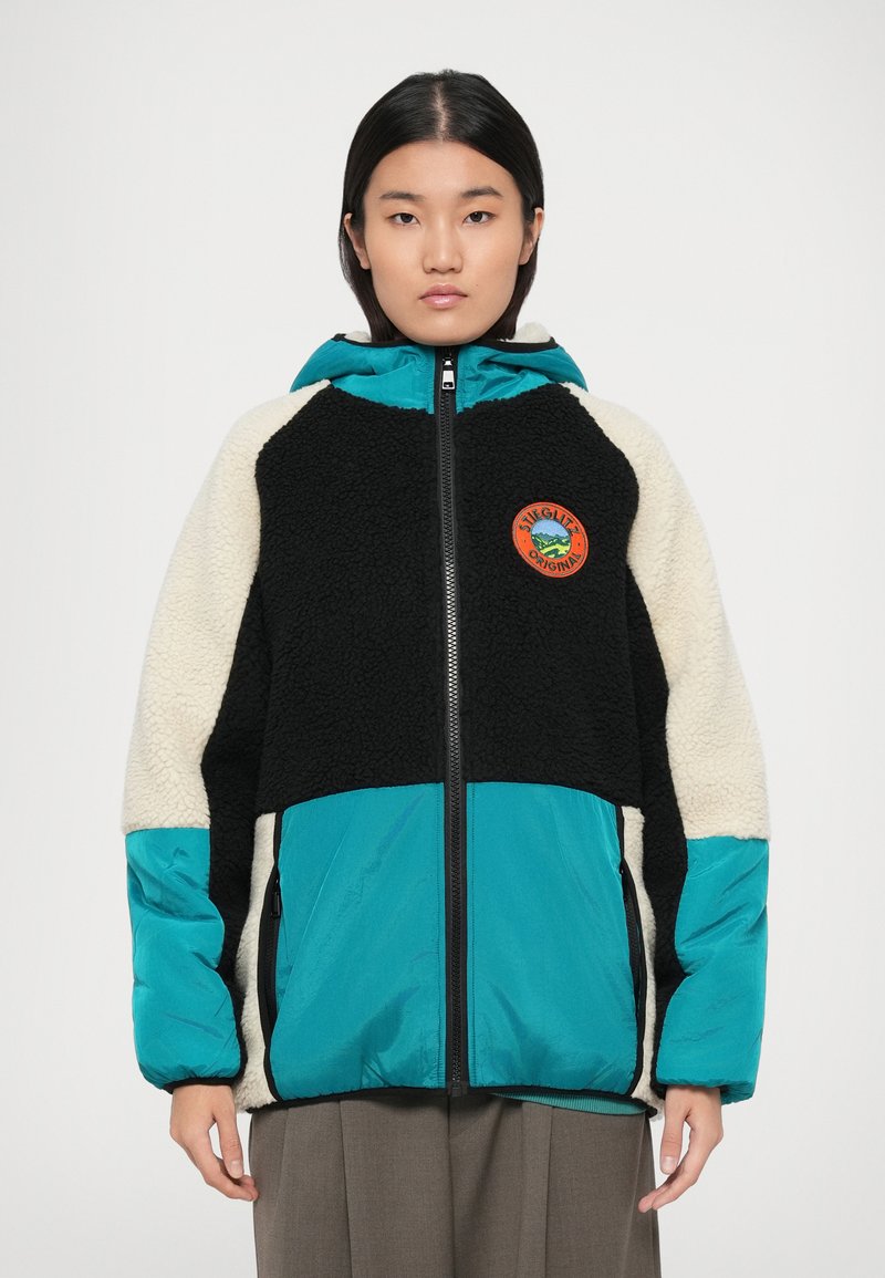 Jachetă colorată cu fleece negru, mâneci crem și baza turcoaz. Include un logo rotund sub formă de patch și fermoare laterale. Se remarcă prin design.
