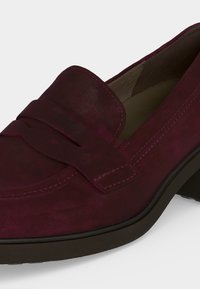 Mocassin en daim bordeaux foncé avec un petit talon, une bride décorative sur le dessus et des coutures visibles le long des bords.