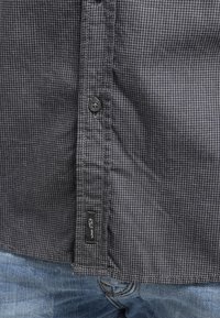 Musta- ja harmaaruutukuvioinen paita, valmistettu kudotusta kankaasta, jossa on napitus ja kudottu etiketti helman kohdalla, yhdistetty sinisiin farkkuihin.