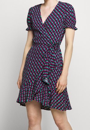 Frau trägt kurzärmeliges Wickelkleid mit pinkem, türkisfarbenem und schwarzem geometrischem Muster und Rüschensaum vor schlichtem Hintergrund.