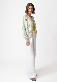 Stickad cardigan med blommönster i grönt, orange och gult. Kombinerad med en gul topp och vita vida byxor. Avslappnad look.