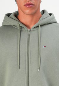 Hombre vistiendo una sudadera con cremallera de color gris claro, con cordones y un pequeño logo rojo, blanco y azul en el lado izquierdo del pecho.