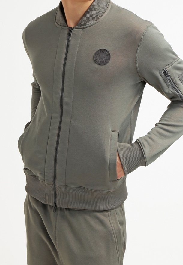 Giacca bomber con zip in verde oliva, realizzata in materiale morbido. Presenta polsini a coste, una toppa rotonda con logo e tasche laterali.