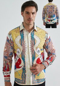 Un homme portant une chemise à motifs colorés featuring des soleils, des étoiles, des écus et des designs ornés, montré de face et de dos.