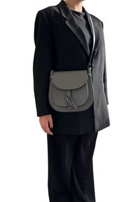 Persona che indossa un blazer nero e pantaloni, con una borsa a tracolla grigia con un dettaglio a frange sulla spalla.