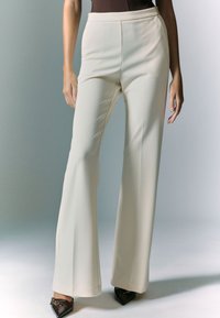 Pantaloni color crema, a vita alta, con una forma svasata, caratterizzati da una texture liscia e dettagli di cuciture verticali, abbinati a scarpe nere a punta.
