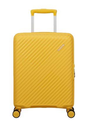 Valigia rigida gialla con scanalature diagonali, manico estendibile, quattro ruote e logo American Tourister sulla parte anteriore.