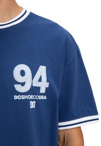 Modrá bavlnená tričko s bielym rebrovaným golierom a okrajmi rukávov. Na hrudi je veľká biela grafika "94" a text "DCSHOECOUSA".