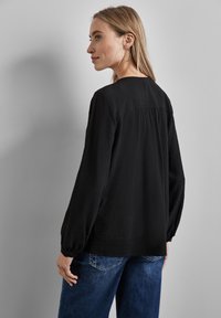 Blusa nera con texture, maniche lunghe e polsi arricciati, con un delicato dettaglio sul retro. Abbinata a jeans in denim blu.