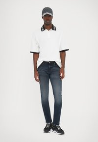 Λευκό polo μπλουζάκι με μαύρες λεπτομέρειες, σκούρο τζην παντελόνι και μαύρα αθλητικά παπούτσια με λευκή επιγραφή. Απλό φόντο, μίνιμαλ σχεδιαστικά στοιχεία.