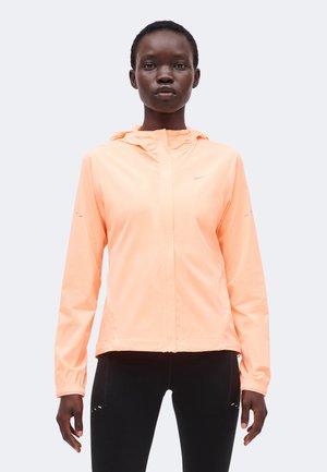 Femme portant une veste à capuche zippée couleur pêche claire et un legging noir, debout, les bras détendus, sur un fond clair uni.