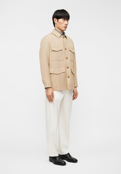 Μπεζ πουκάμισο με κουμπιά, τέσσερις εξωτερικές τσέπες, ελαφριά υφή και γιακά, συνδυασμένο με λευκό παντελόνι και μαύρα παπούτσια.