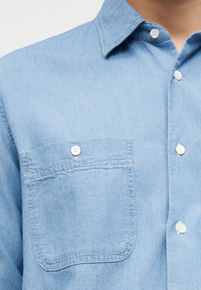 Ανοιχτό γαλάζιο τζιν πουκάμισο με κουμπιά, με τσέπη στο στήθος που κλείνει με λευκό κουμπί, με γιακά ορατό, φορεμένο από ένα άτομο.