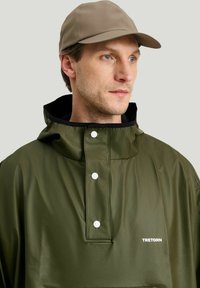 Oliivinvihreä vedenpitävä takki, jossa on korkea kaulus, nepparit ja mustalla vuorattu huppu; vaaleanbeige lippalakki; logo rinnassa.