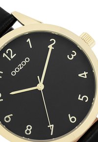 Cadran noir avec index horaires dorés, aiguilles dorées et boîtier doré. Bracelet en cuir noir avec détail de couture. Logo de la marque affiché.