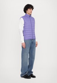 Colete puffy lilás claro com gola alta e fecho de zip, usado sobre uma camisa branca de manga longa, combinado com jeans azuis de perna larga e sapatos pretos.