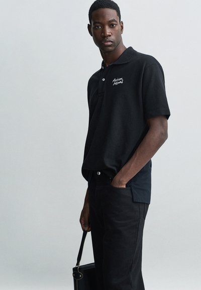 Polo shirt noir à manches courtes, arborant un petit logo blanc brodé sur la poitrine gauche. Associé à un pantalon noir et un sac noir.
