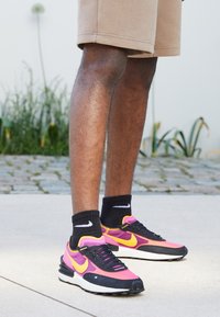 Černé a růžové sportovní boty se žlutými akcenty, s texturovaným svrškem a bílou podrážkou, nošené s černými ponožkami na betonovém povrchu.