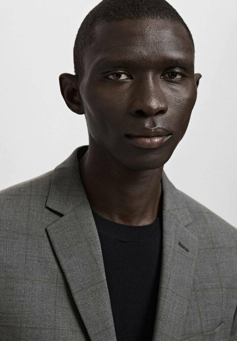 Grauer karierten Blazer über einem schwarzen Strickshirt. Glatte Textur mit strukturierten Revers und minimalistischen Details. Nahaufnahme-Porträt.