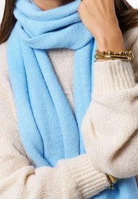 Lichtblauwe wollen sjaal gedrapeerd om een lichtbeige gebreide trui met ribbelmanchetten, met gouden armbanden om de pols.