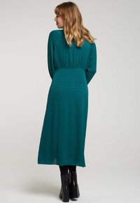 Robe bleu sarcelle mi-longue avec un motif texturé, des manches longues, une taille élastique et une silhouette fluide. Associée à des bottines noires.