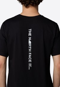 Sort bomulds T-shirt med en lodret hvid logo-print på ryggen, med teksten "THE NORTH FACE" og et buet grafisk element.