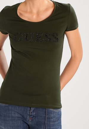 T-shirt cintré vert olive à manches courtes, arborant un logo "GUESS" en strass noir sur la poitrine. Texture lisse, col rond.