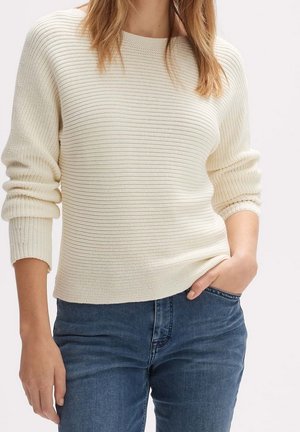 Femme portant un pull en maille côtelée crème à manches longues et un jean bleu, main dans la poche, cadrée en dessous du cou.
