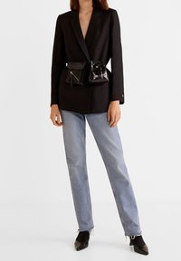 Blazer nero su misura con revers, abbinato a jeans dritti azzurri chiari. Presenta due piccole borse nere attaccate in vita. Scarpe nere con punta appuntita.