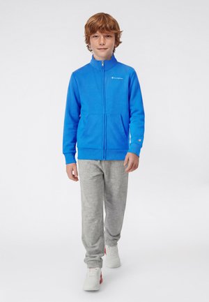 Chłopiec o rudych włosach idący naprzód, ubrany w niebieską kurtkę na zamek, szare dresy i białe trampki na białym tle.