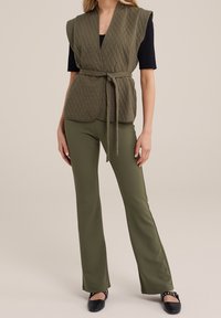 Oliivinvihreä tikattu liivi solmivyöllä, jossa on v-kaulus ja hartiakappaleet. Yhdistetty vastaaviin oliivinvihreisiin levenevät housut ja mustat kengät.