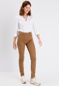 Pantalons skinny en mélange de coton marron, chemise blanche à boutons, chaussures de sport crème et beige, look décontracté, présentés sur une figure debout.