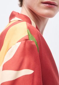 Blusa a motivo rosso e giallo con maniche corte, con accenti verdi. Tessuto liscio e leggero con una lucentezza morbida.