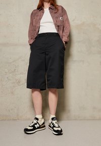Osoba v černých kraťasech po kolena, béžových ponožkách, černobílých teniskách New Balance, bílém triku a mauve košili se zapínáním proti jednoduché zdi.