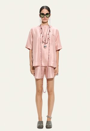 Kobieta ubrana w bladoróżowy satynowy krótki top i szorty, długie naszyjniki z koralików, okrągłe okulary przeciwsłoneczne, szare wsuwane buty oraz bransoletkę na kostkę, stoi na białym tle.