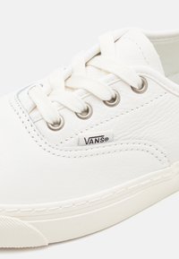 Baskets Vans blanches en cuir texturé, avec des lacets blancs, des œillets en métal et un petit logo noir Vans sur le côté.