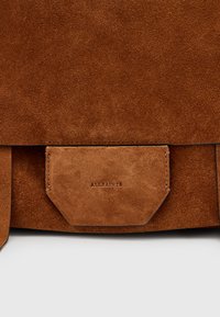 Borsa in suede marrone con texture liscia, dotata di chiusura a pattina e tasca frontale. Un branding sottile è impresso sulla tasca.