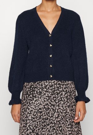 Kobieta nosząca granatowy granatowy kardigan z dekoltem w serek i guzikami w kształcie serc oraz czarno-beżową spódnicę w wzory.