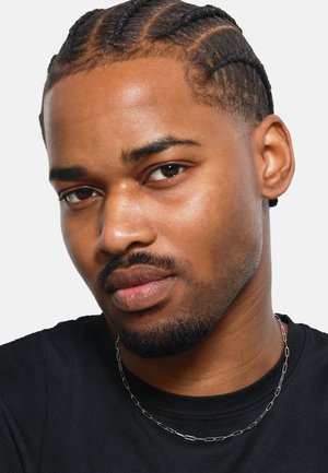 Headshot of a person with braided hair, wearing a black shirt and a thin silver chain necklace, featuring smooth skin and neutral expression.