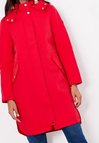 Parka impermeable roja con cierre de botones a presión, dos bolsillos laterales y capucha con cierres a presión. Textura lisa y diseño hasta la rodilla.