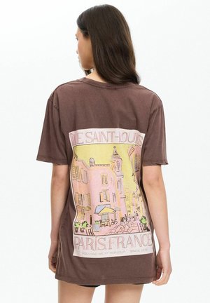 Mujer con cabello largo y oscuro que lleva una camiseta marrón con una ilustración en rosa y amarillo de una escena callejera de París en la espalda.