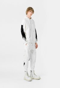 Hoodie blanco con detalles de alas negras, combinado con pantalones blancos que presentan un diseño de plumas. Botas blancas con cordones y suelas gruesas.