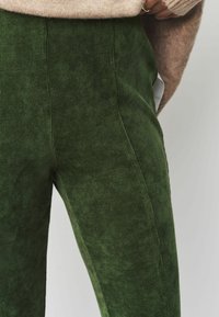 Pantalon en velours côtelé vert avec une finition texturée, présentant des côtes verticales et une coupe ajustée. Poches latérales visibles à la taille.