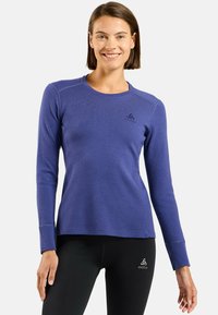 Femme aux cheveux bruns courts souriant, portant un haut de sport bleu à manches longues et un legging noir avec des logos Odlo sur les deux vêtements.