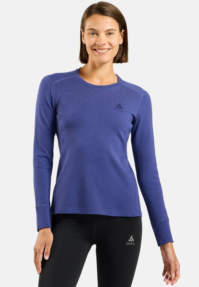 Femme aux cheveux bruns courts souriant, portant un haut de sport bleu à manches longues et un legging noir avec des logos Odlo sur les deux vêtements.