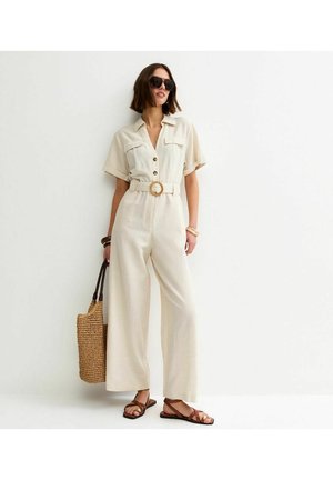 Mujer con un mono color crema con cinturón, gafas de sol oscuras, sandalias marrones y llevando un bolso de mano de paja tejido, parada contra una pared blanca.