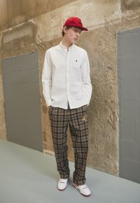 Chemise blanche à boutonnage, pantalon à carreaux marron avec des coutures dorées, casquette rouge et chaussures blanches. Tissu doux avec une coupe décontractée, style casual.