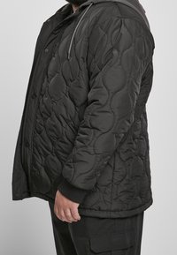 Homme portant une veste matelassée noire avec capuche et un pantalon cargo noir, debout de profil sur un fond uni.