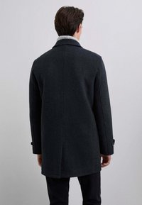 Abrigo de lana oscura con un diseño estructurado, un cuello gris claro en contraste, mangas largas y puños abotonados. Estilo de corte recto.